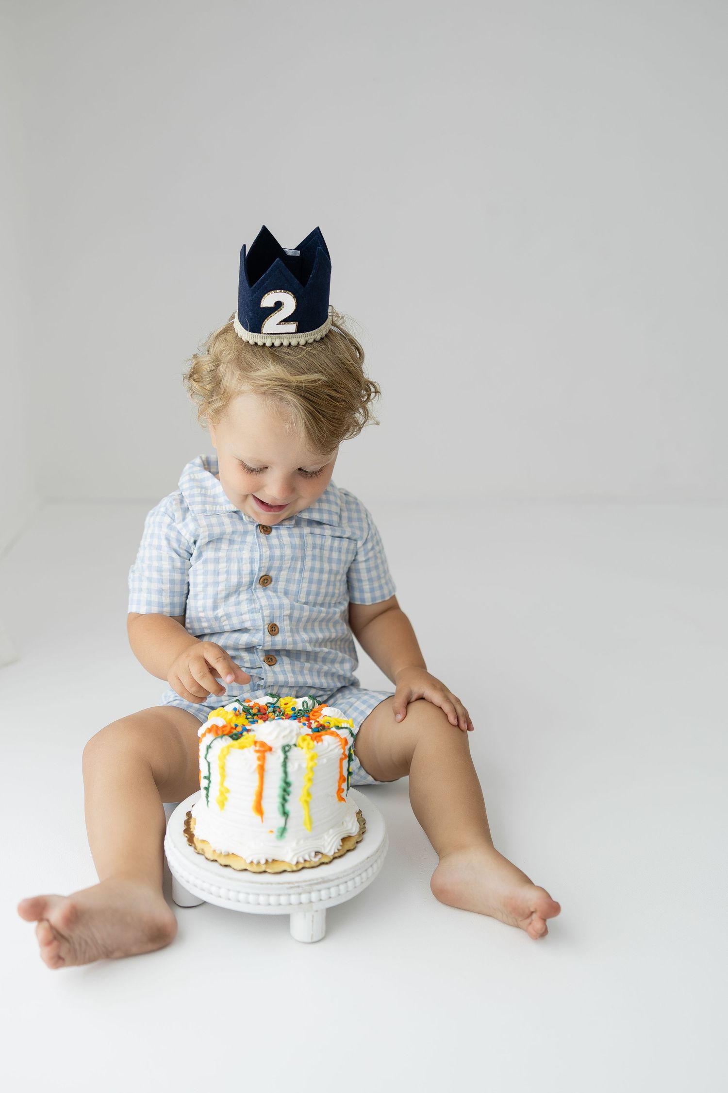 Toddler boy wearing a number 2 crown sits on white floor smiling at colorful birthday smash cake.