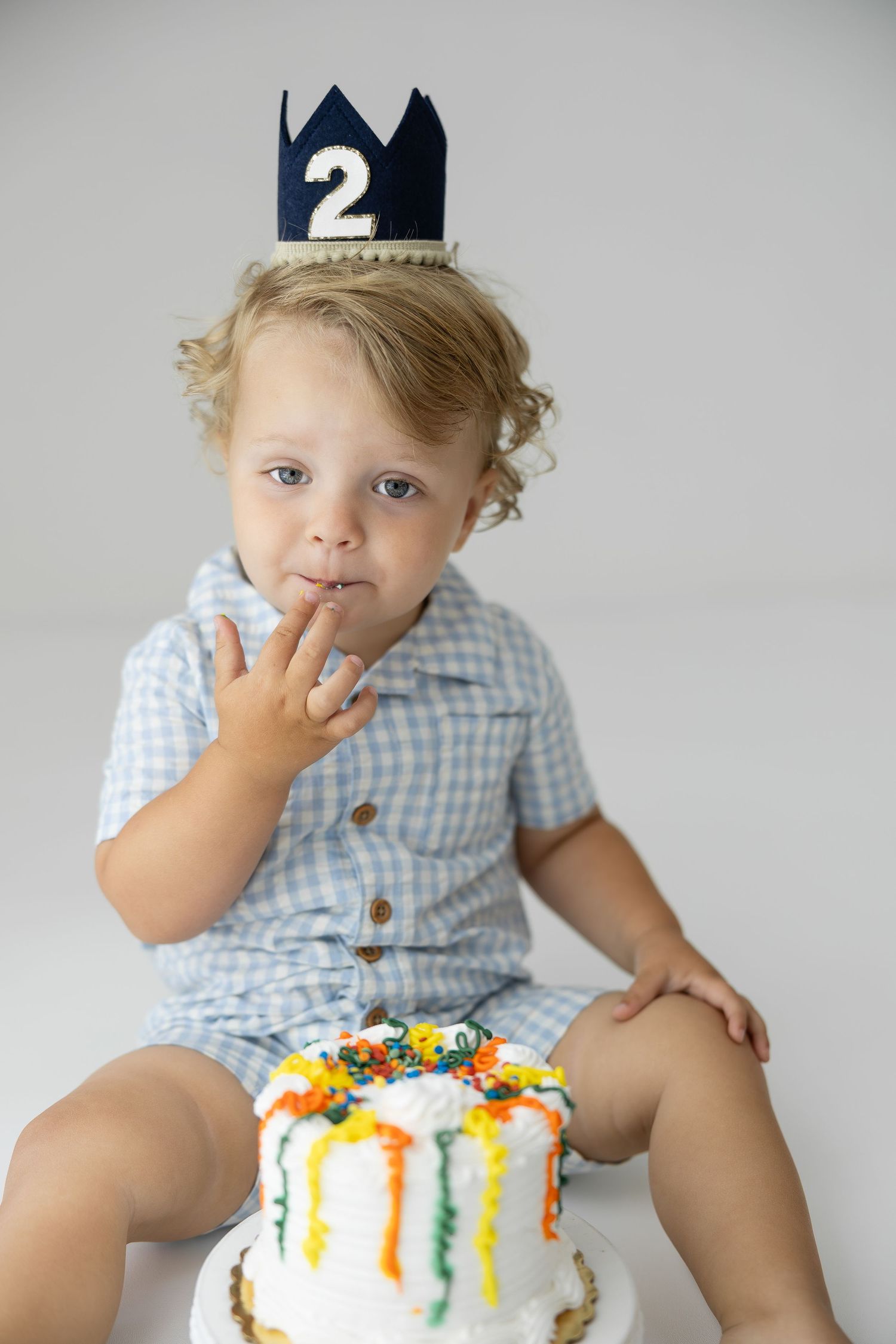 Toddler boy wearing a number 2 crown licks frosting from his finger beside a colorful smash cake on his 2nd birthday.