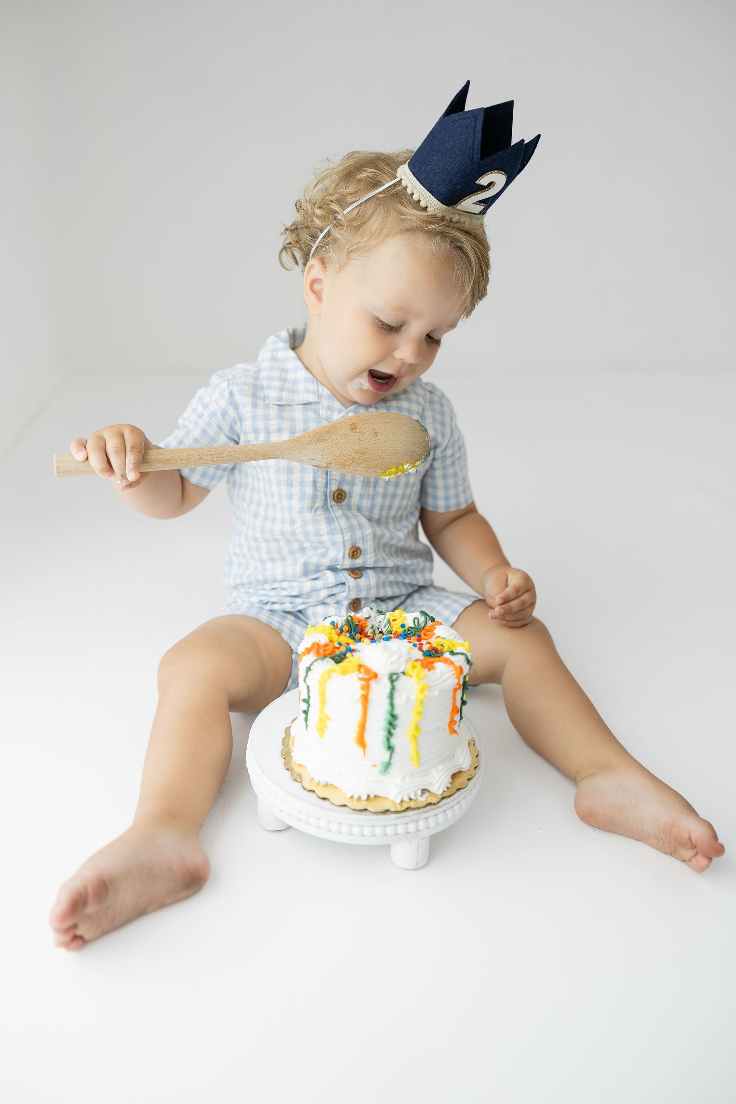 Toddler in blue outfit and paper crown holds wooden spoon over colorful birthday cake on white background.