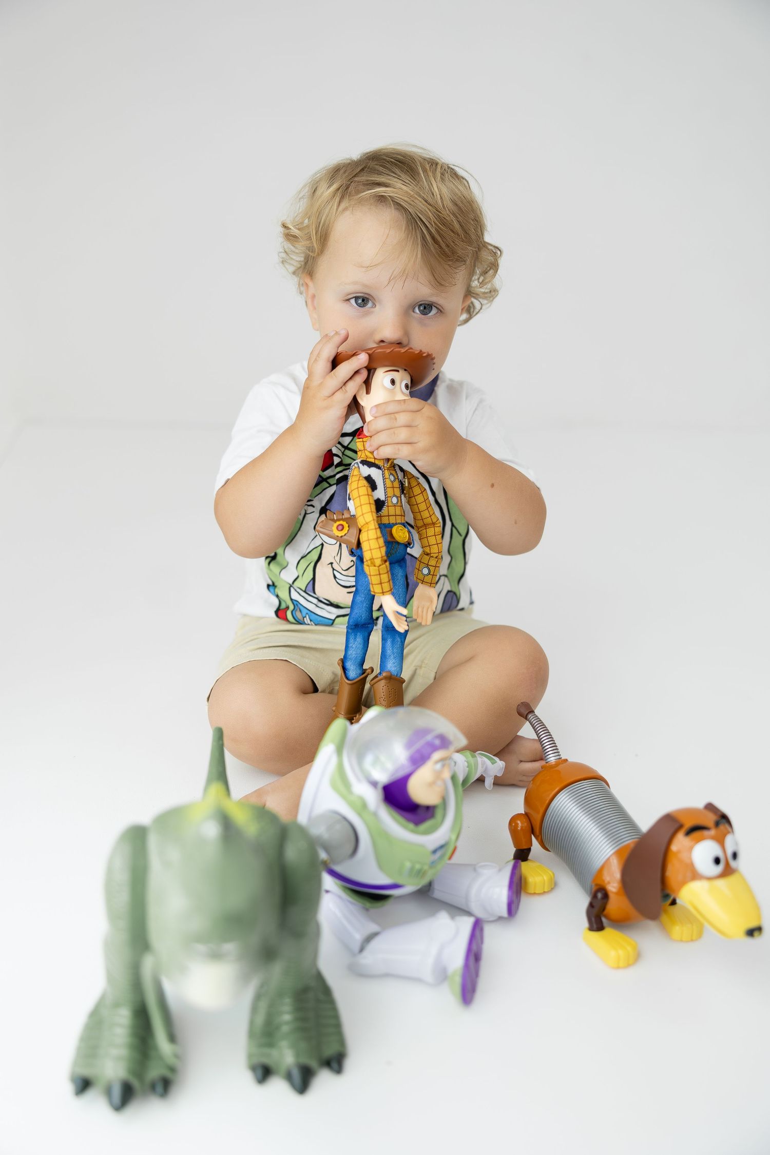 Toddler boy chewing Woody doll surrounded by Toy Story toys including Rex, Buzz Lightyear, and Slinky Dog.