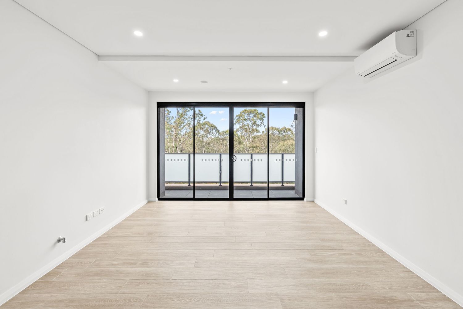 Bright empty apartment room with large sliding glass doors opening to a balcony with garden views.