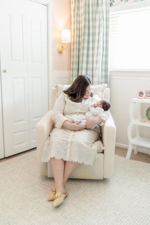 A mother in a cream dress sits in a glider chair cradling her newborn baby in a soft, pastel nursery room.
