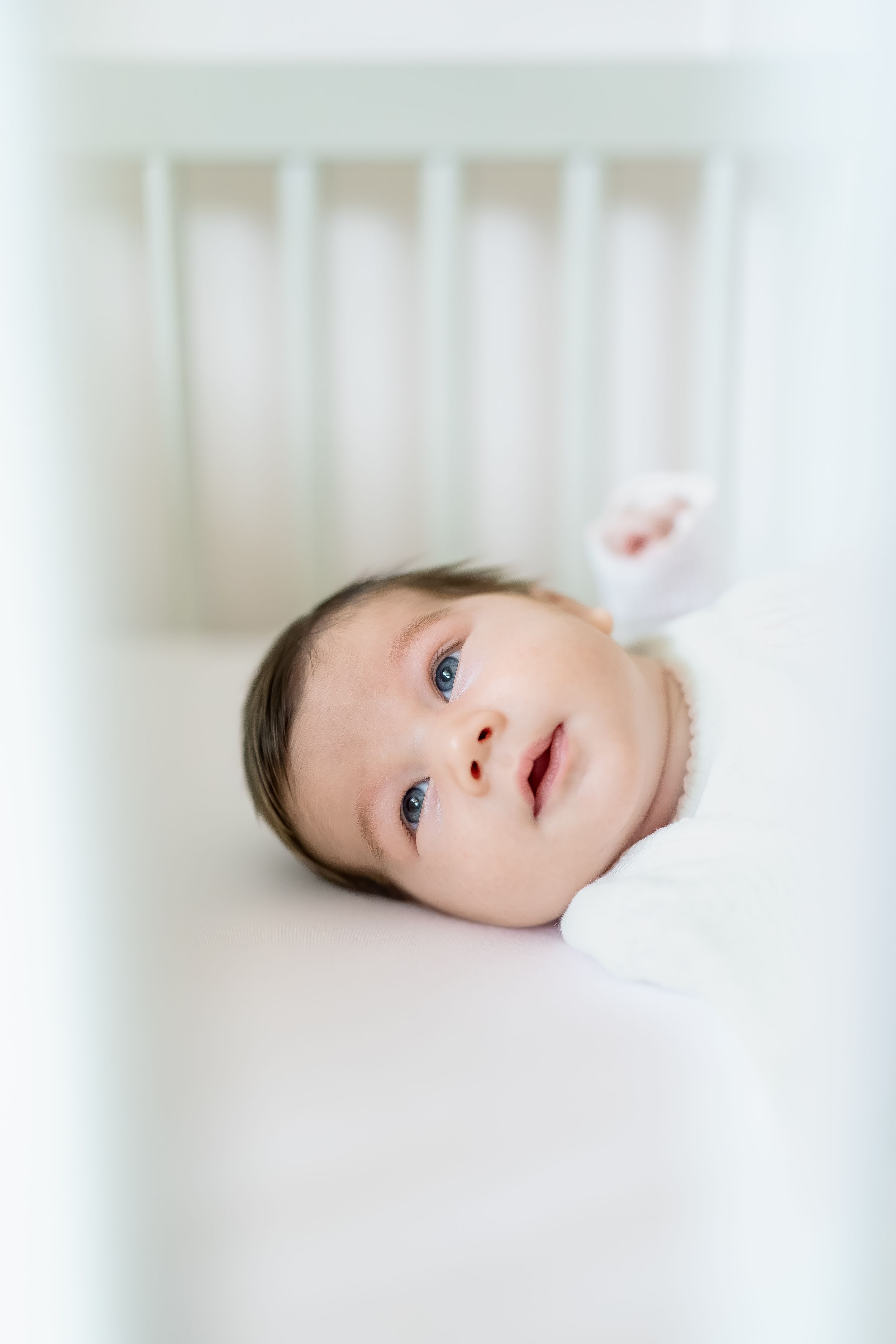 Newborn baby with blue eyes lying awake in a white crib, wrapped in a soft white swaddle blanket.