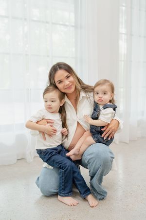 A smiling mother crouches on a white floor, hugging her two young toddlers dressed in jeans and white tops.