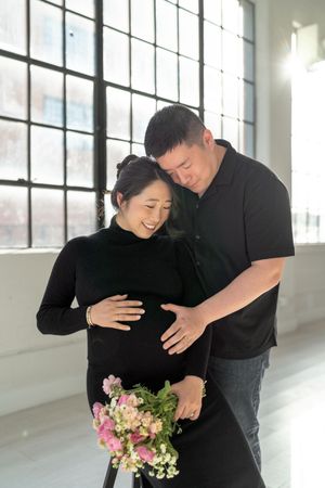 Smiling pregnant woman in black holding flowers, embraced by man in black polo, maternity photo session.