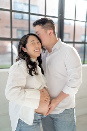 A happy pregnant Asian couple in white shirts share a tender kiss, with the man gently holding her baby bump.