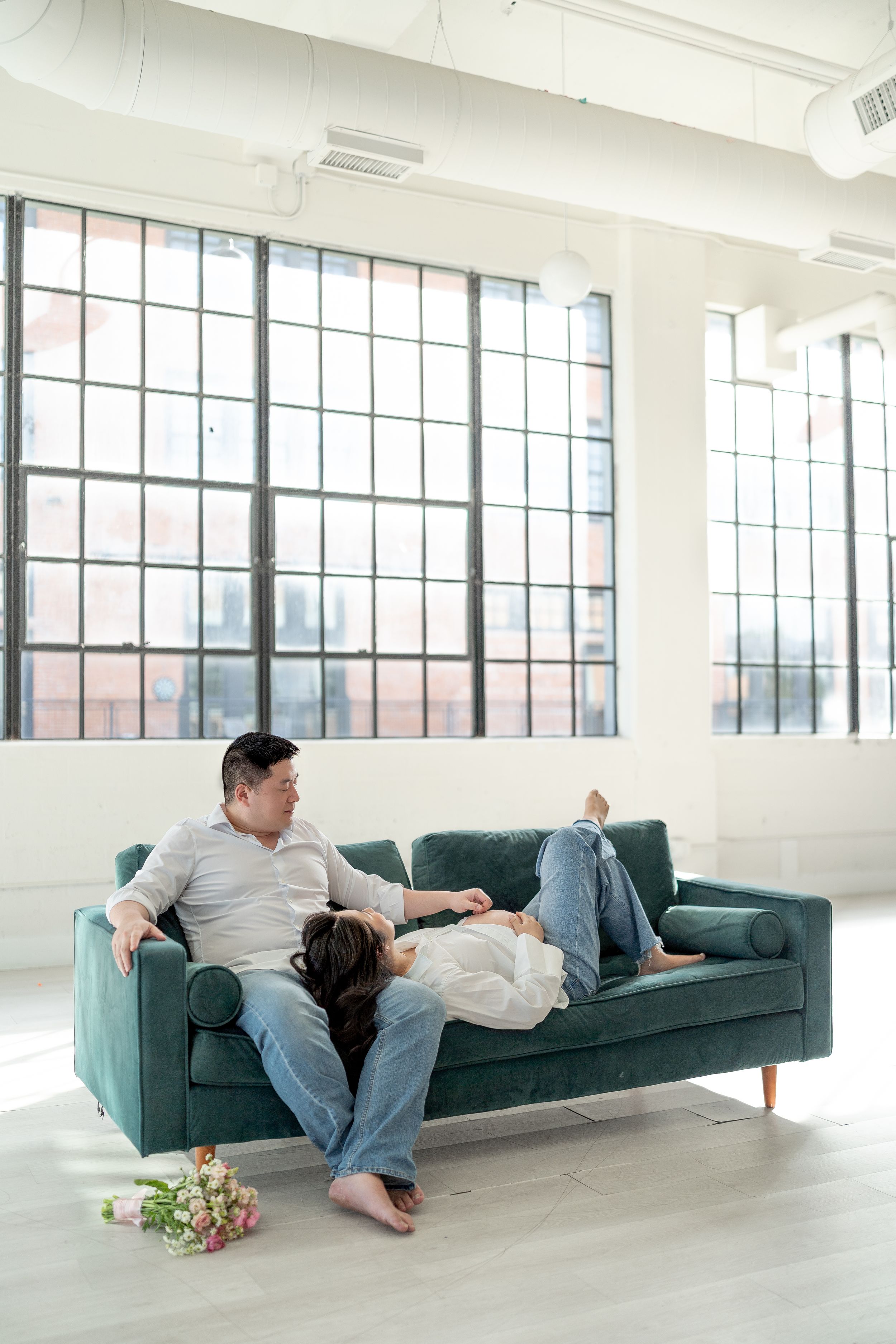 Couple relaxing on a teal velvet sofa in a bright loft, woman lying across man's lap near a floral bouquet.