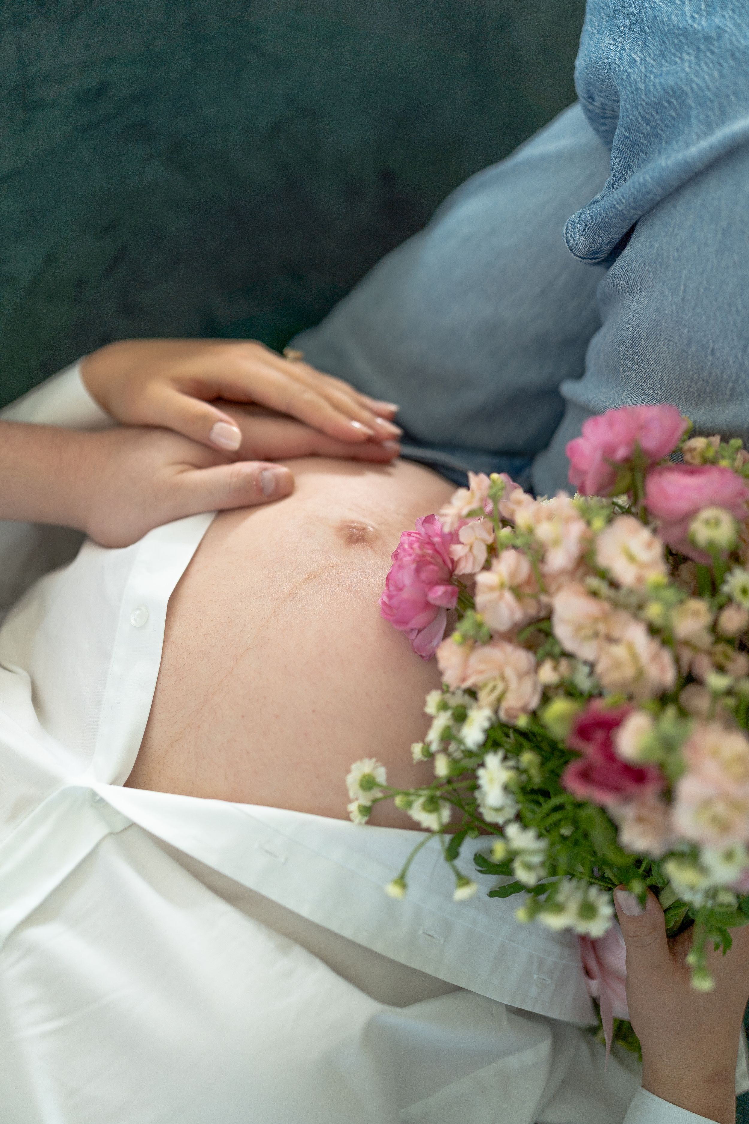 Pregnant woman resting on sofa holding pink flower bouquet on bare belly, maternity photography concept.