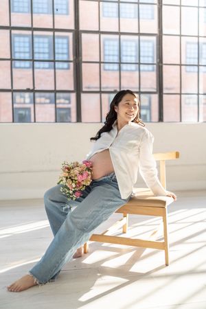 Smiling pregnant woman in white shirt and jeans holding flowers, sitting on wooden chair in sunlit loft studio.