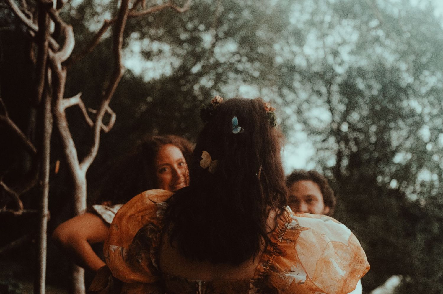 Three women in floral dresses share a warm embrace outdoors surrounded by trees in soft natural light.