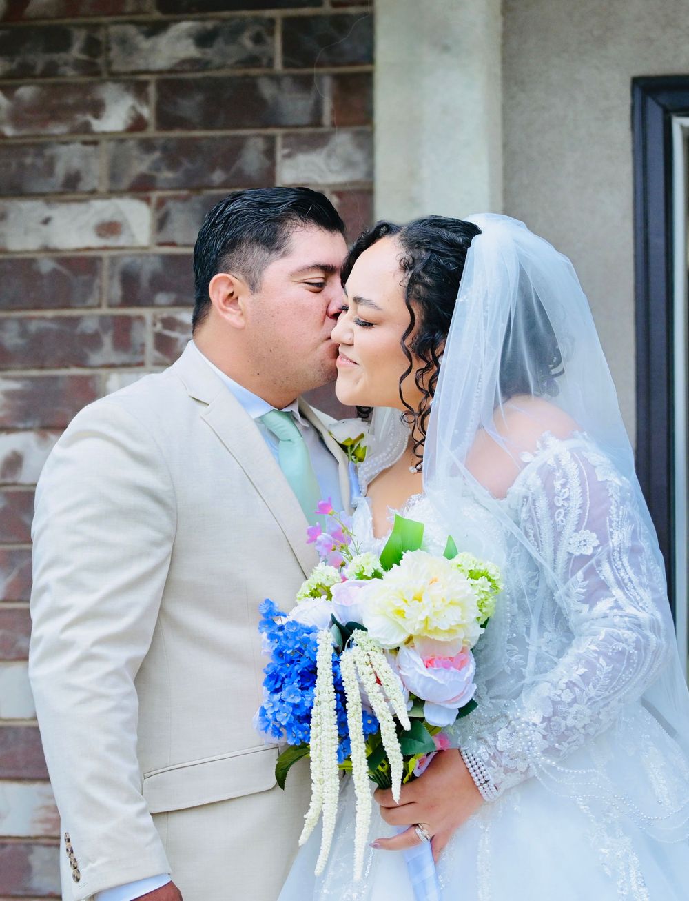 Bride and groom sharing a kiss on their wedding day, bride holding a colorful bouquet of blue hydrangeas and white flowers.