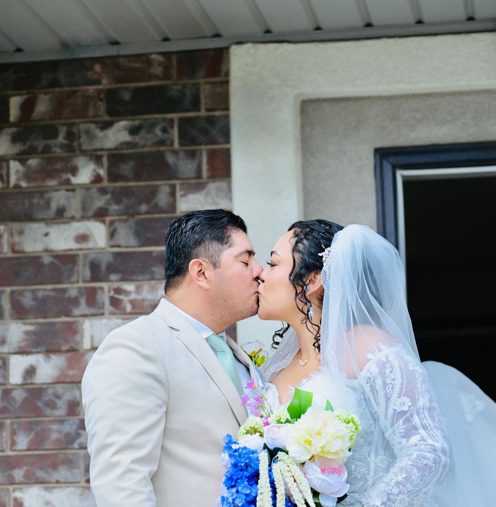 Wedding couple sharing first kiss in front of brick building, bride in lace gown with veil holding colorful bouquet.