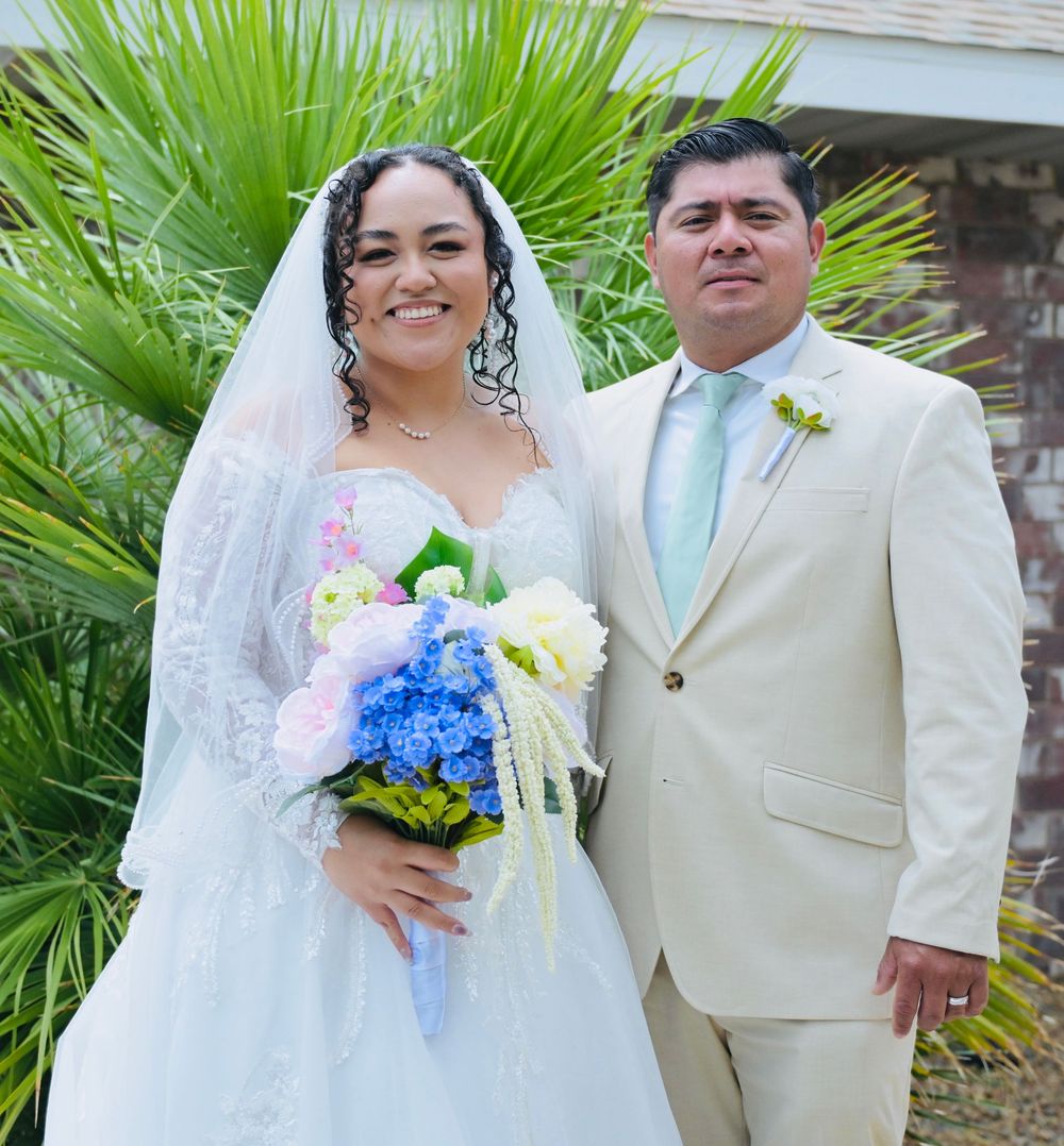 Smiling bride and groom posing together outdoors on their wedding day, bride holding a vibrant blue and white bouquet.