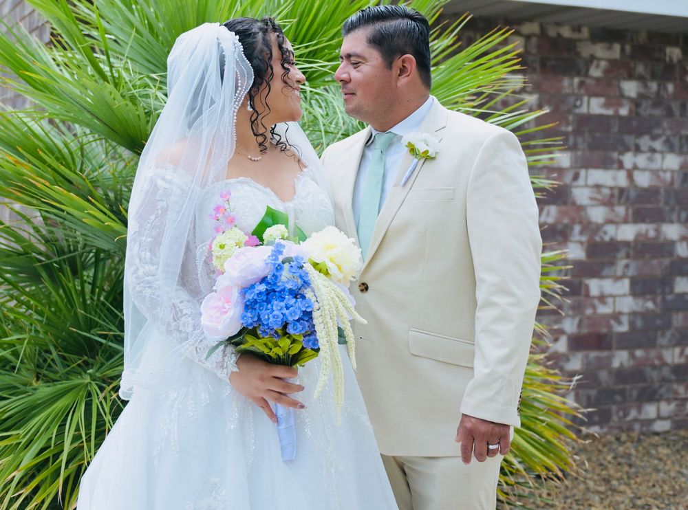 Wedding couple gazing at each other outdoors near palm trees, bride in lace gown holding a colorful hydrangea bouquet.
