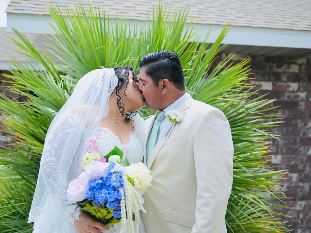 Newlyweds sharing first kiss outdoors, bride with blue floral bouquet, groom in cream suit, tropical palm background.