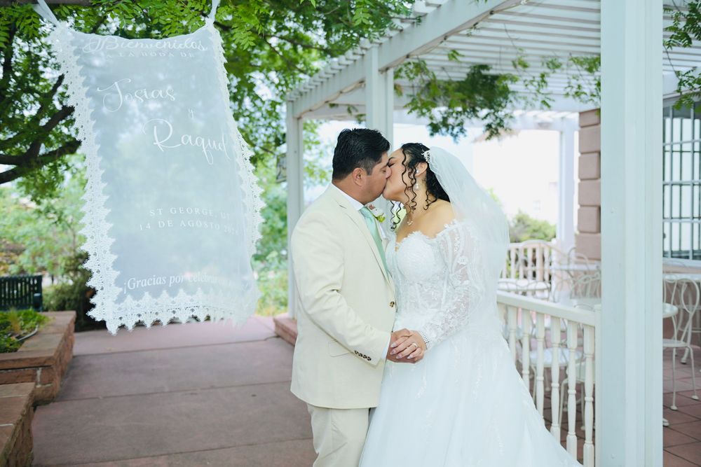 Bride and groom share a romantic kiss under a white gazebo with a decorative wedding sign hanging nearby.