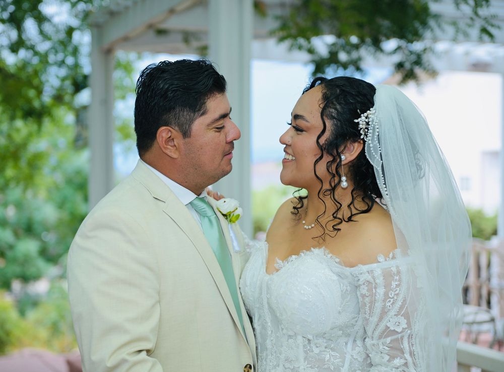 Bride and groom in cream suit share romantic moment under white pergola surrounded by lush greenery.