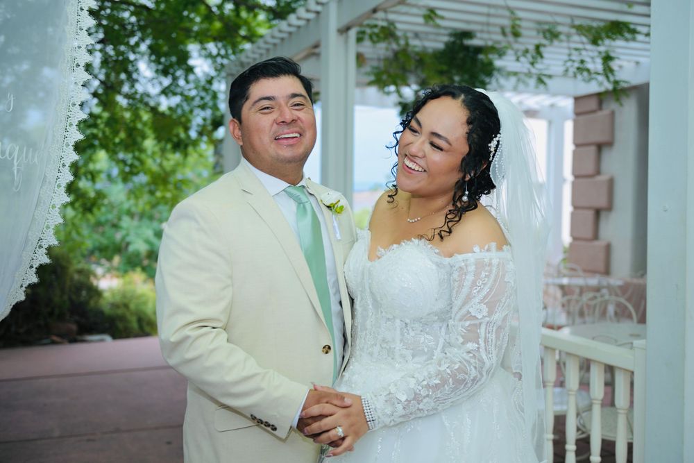 Smiling newlyweds embrace under white pergola, groom in cream suit and bride in off-shoulder lace wedding gown.