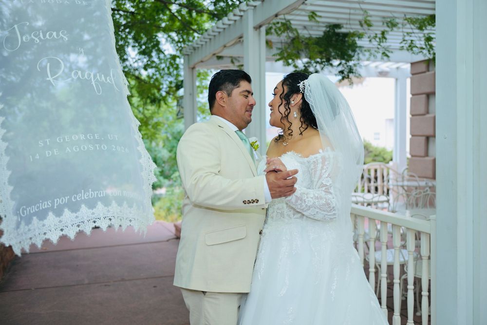 Wedding couple sharing romantic moment on white gazebo porch, bride in lace gown with veil, groom in cream suit.