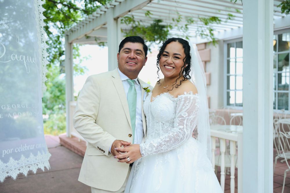 Smiling couple in wedding attire pose under a white gazebo, bride in lace gown and groom in tan suit with mint tie.