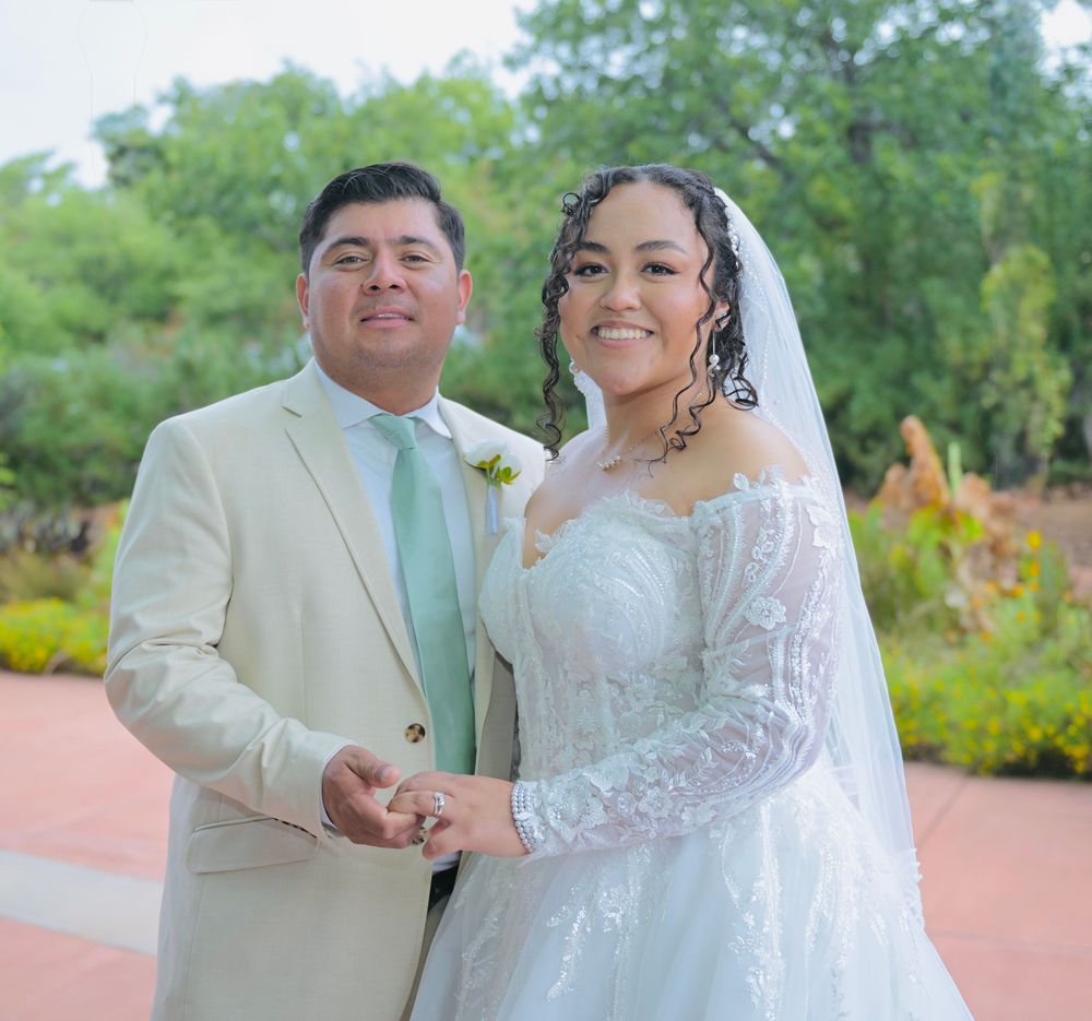 A smiling bride in a white lace gown and veil holds hands with a groom in a beige suit at an outdoor wedding.