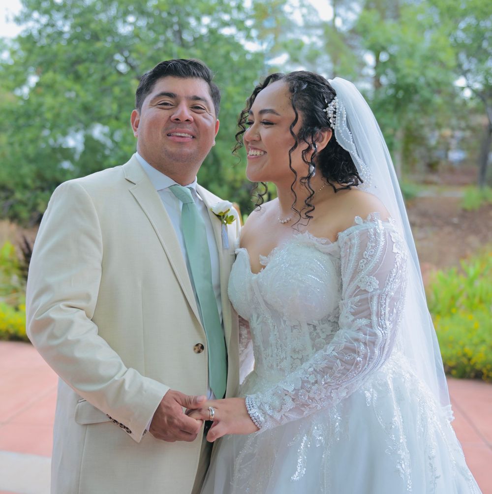 Smiling bride and groom pose outdoors on their wedding day, she in lace gown with veil, he in cream suit with mint tie.