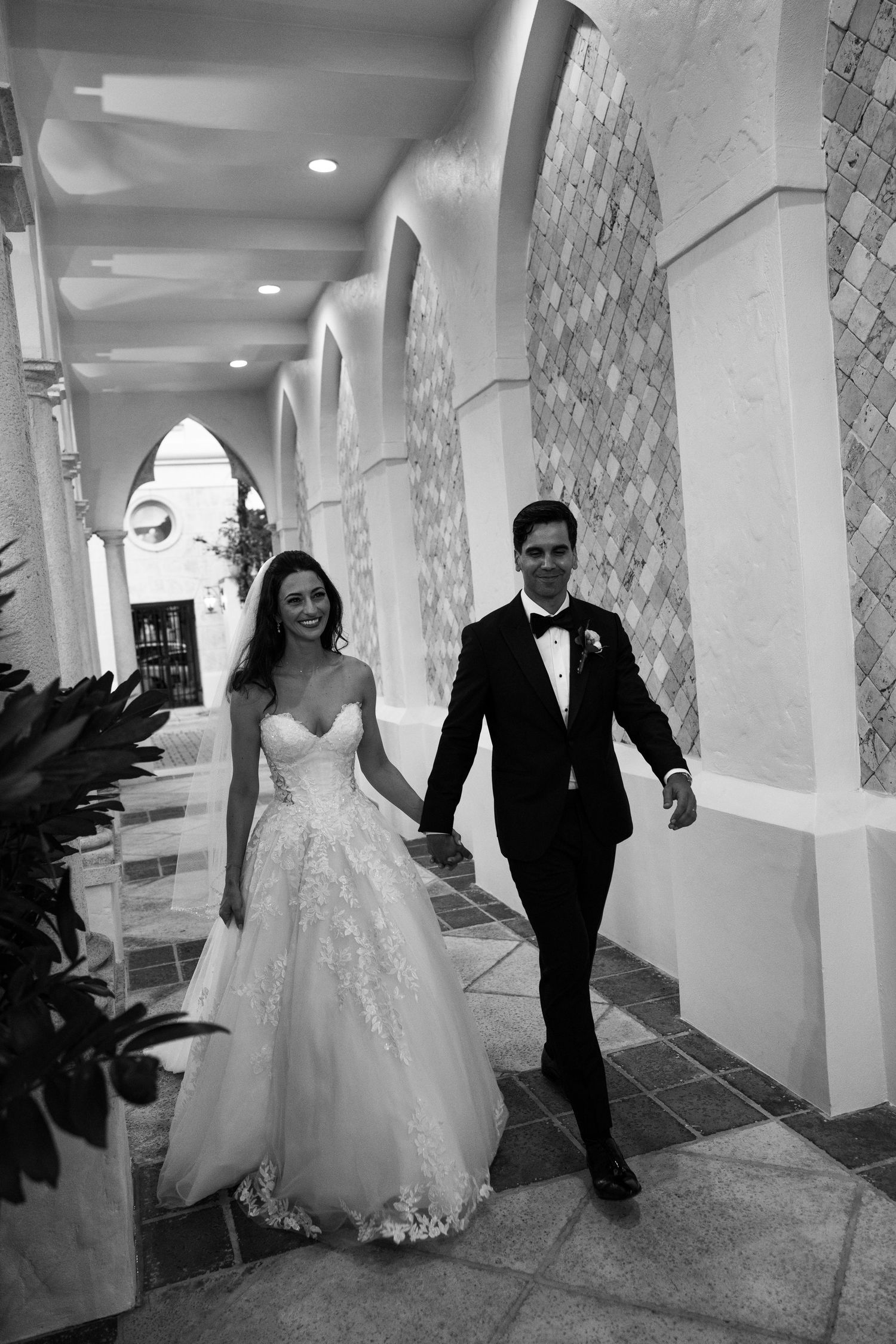Black and white photo of a bride and groom holding hands walking through an elegant arched corridor on their wedding day.