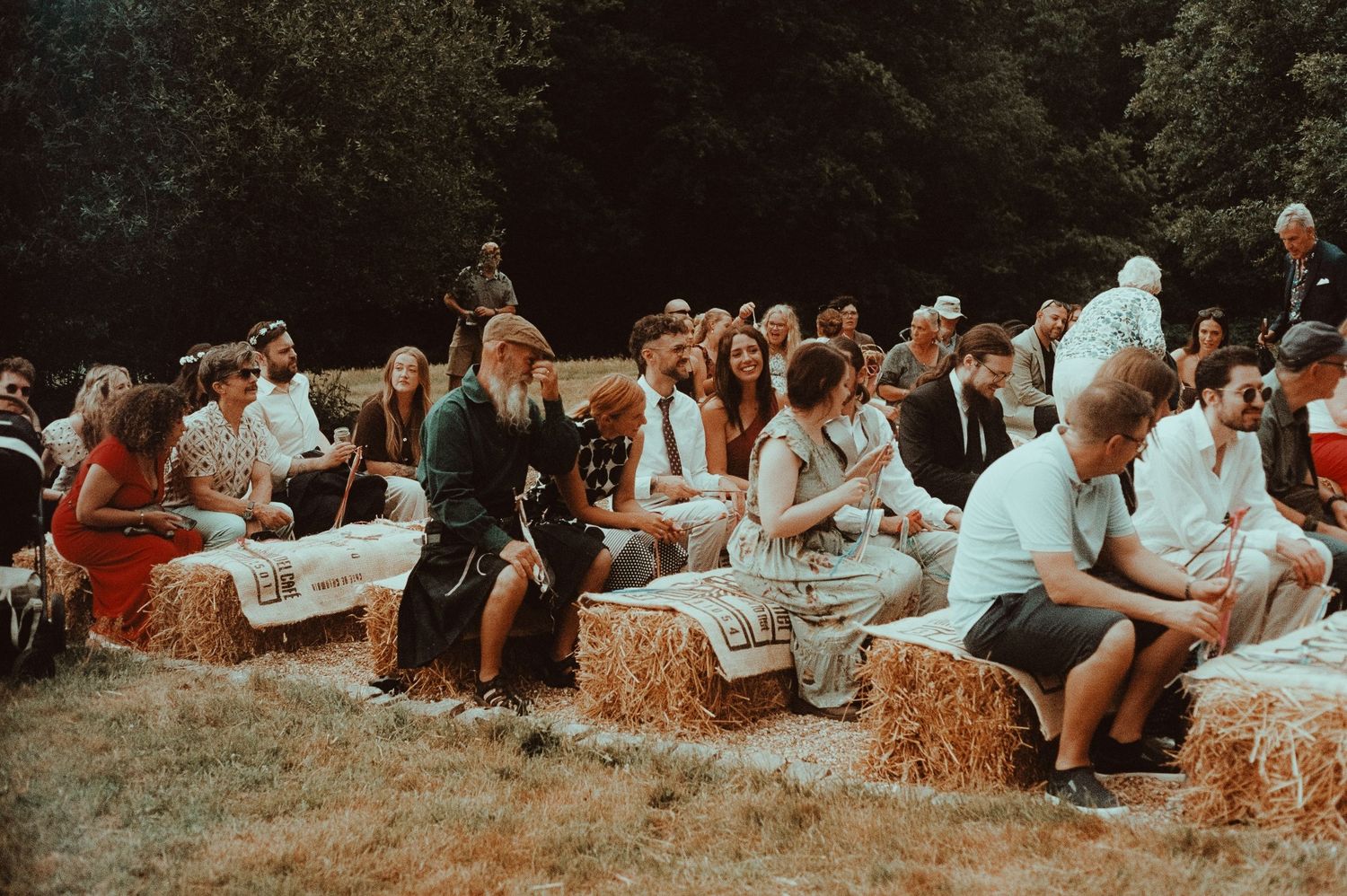 Guests seated on hay bales at an outdoor wedding ceremony surrounded by lush green trees on a sunny day.
