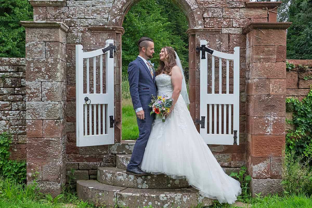 Bride and groom share a romantic moment framed by white garden gates and stone Dalston Hall wedding venue near Carlisle.
