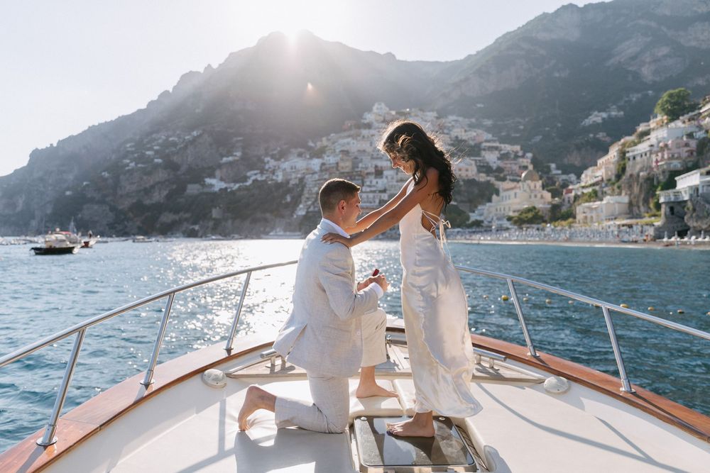 Man proposing on one knee to woman on luxury yacht with Amalfi Coast cliffs and sunlight in background.