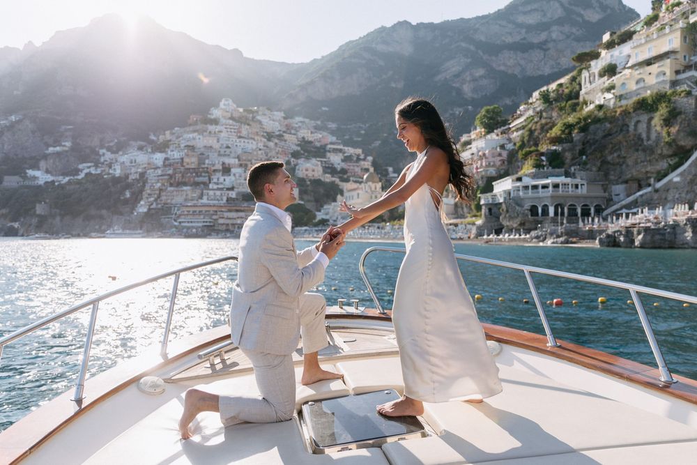 Man proposes on luxury yacht deck with stunning Amalfi Coast backdrop, couple in white attire.