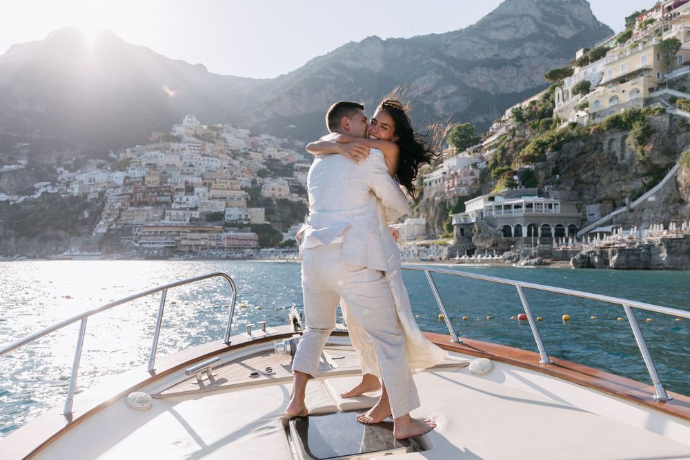 Couple embracing and kissing on a luxury yacht bow with Positano, Amalfi Coast cliffside village in background.
