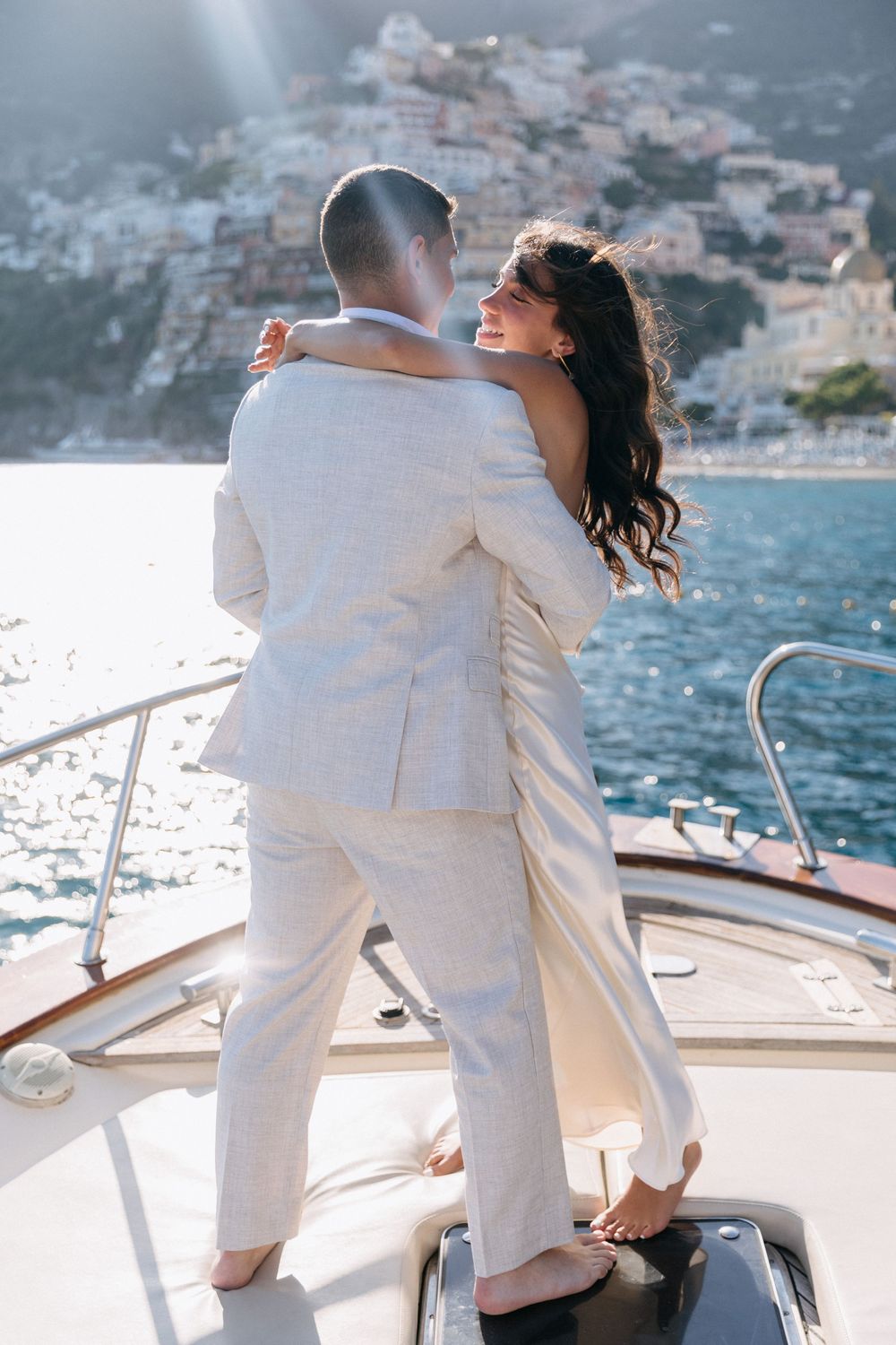 Couple embracing on a yacht bow with Positano, Amalfi Coast in the background on a sunny day.
