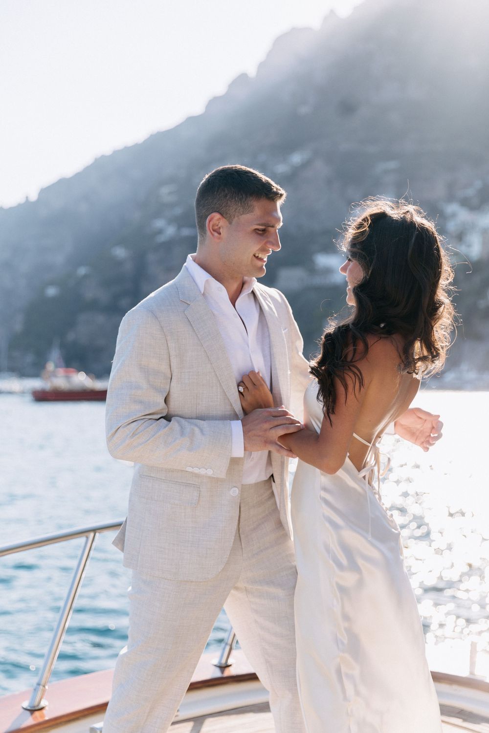 A smiling couple in white wedding attire dance on a yacht with scenic Positano mountains and sparkling water behind them