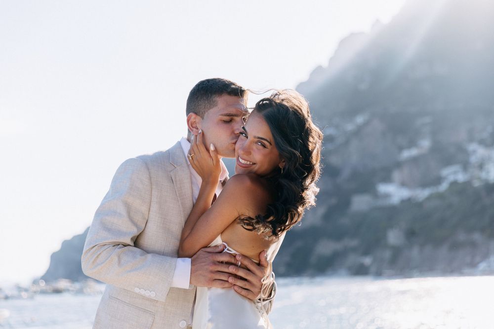 Romantic couple embracing on a sunny coastal cliffside, groom in linen suit kissing bride in white dress.