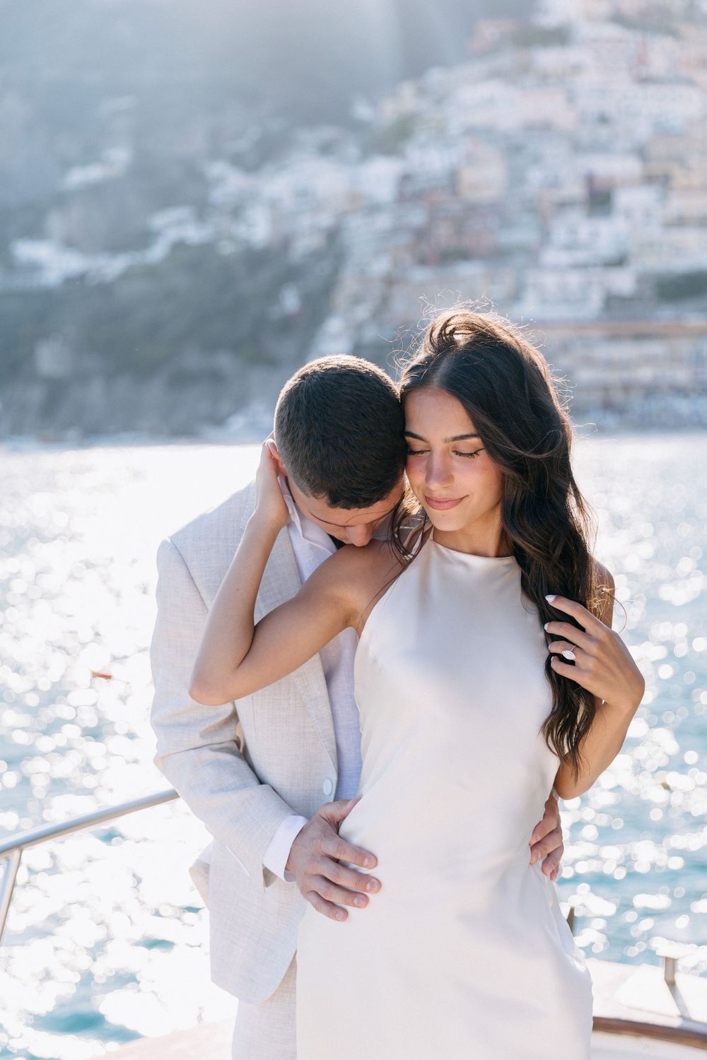 Romantic couple embracing on a yacht with Positano Italy coastal village in the sunlit background.
