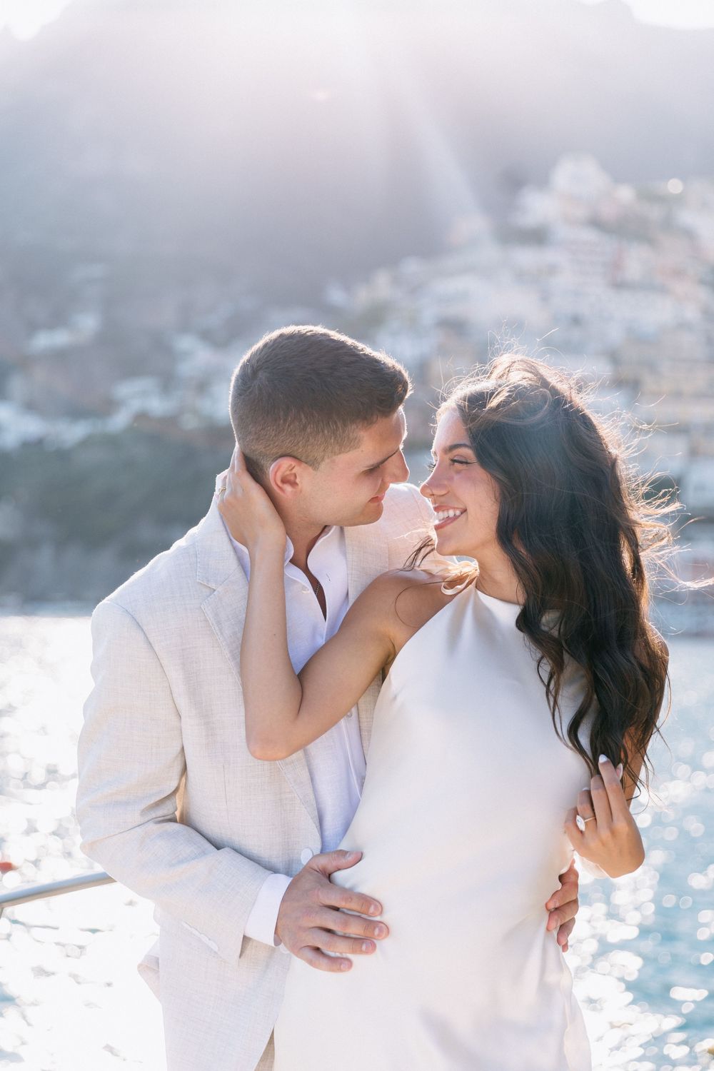 Couple in white attire embracing and smiling at sunset with Positano coastal town in the background.