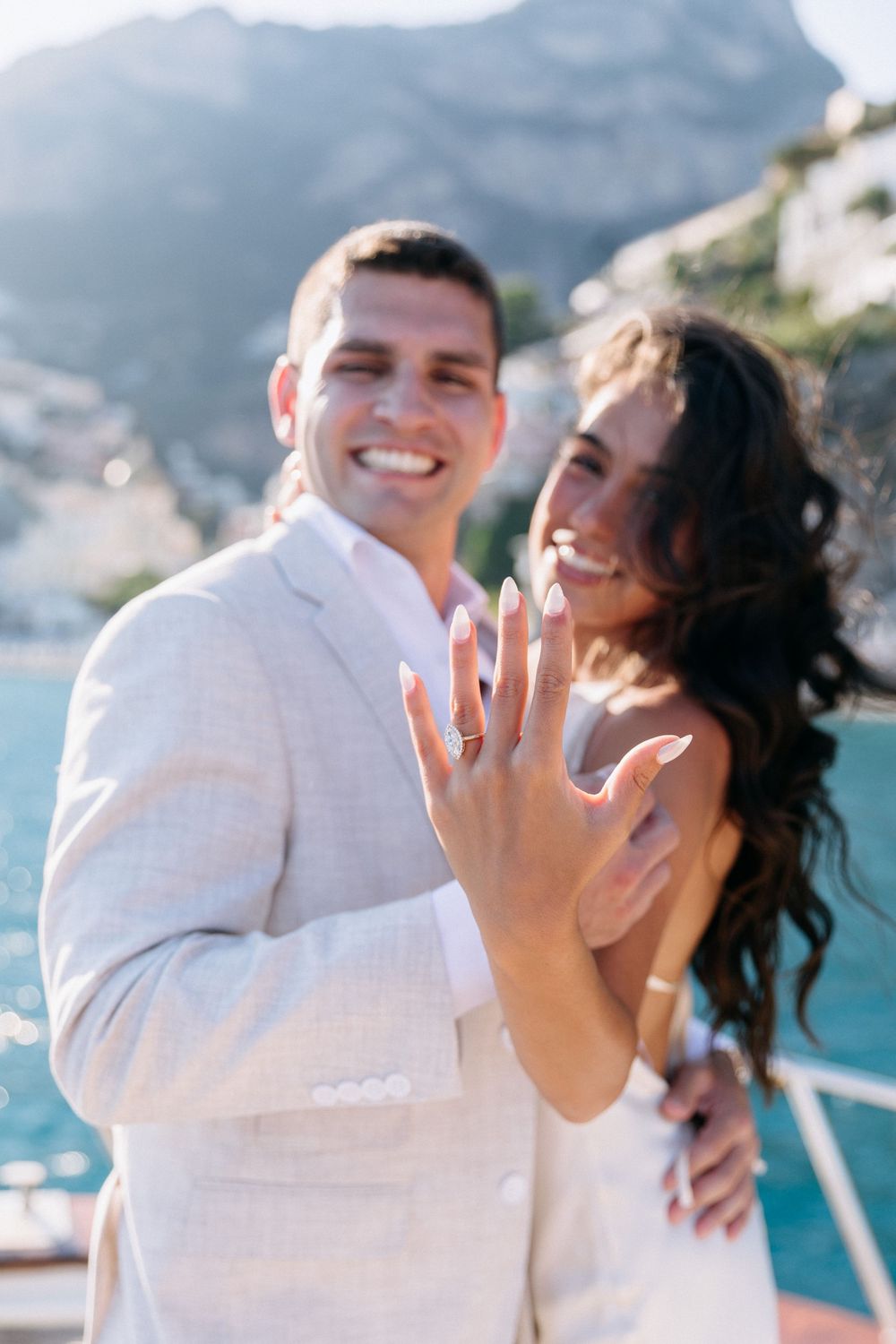 Happy couple on a yacht showing off their engagement rings with scenic coastal Amalfi Coast cliffs in the background.