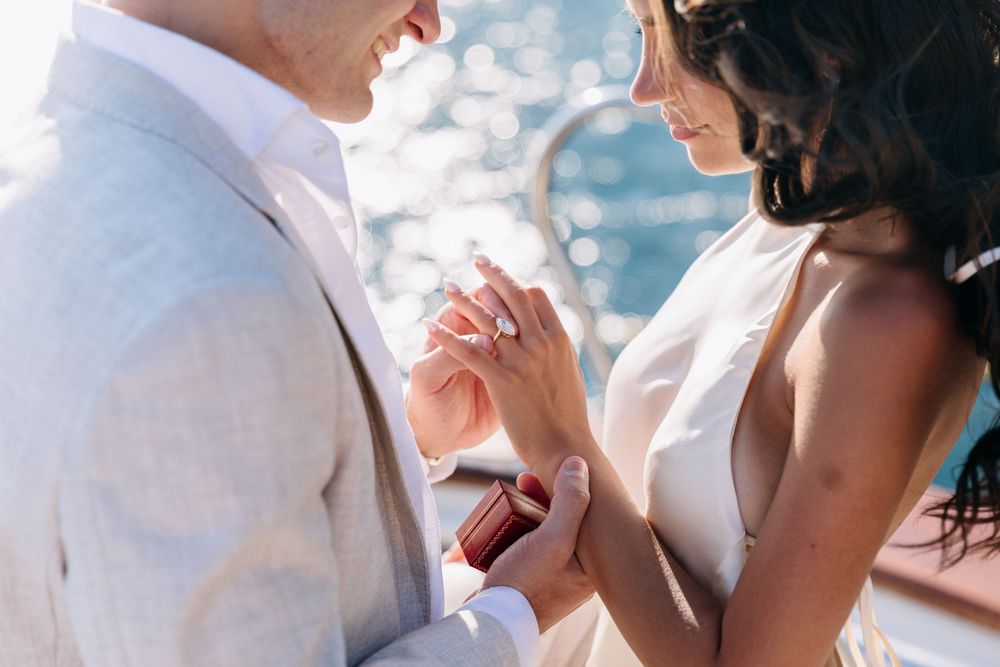 Groom placing a diamond engagement ring on bride's finger during a romantic outdoor wedding ceremony.