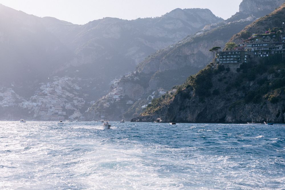Scenic Amalfi Coast view from the sea with boats on blue water and cliffside buildings nestled in rugged mountains.