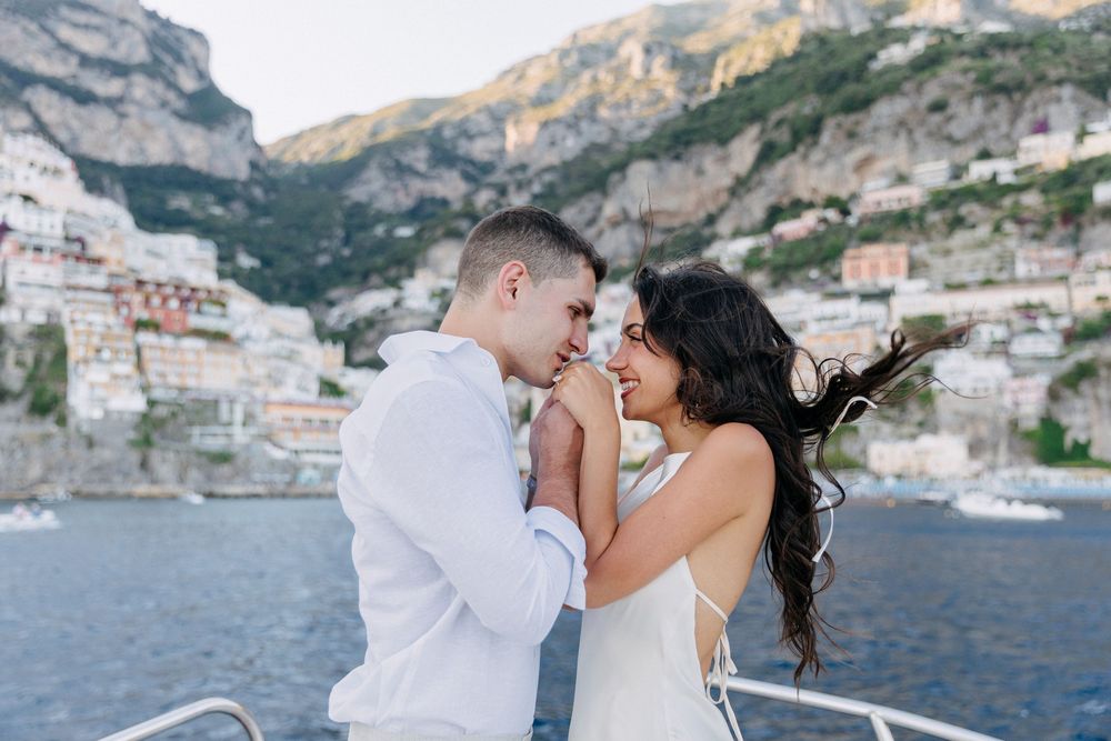 Couple sharing a romantic moment on a yacht with the stunning Amalfi Coast cliffside village in the background.