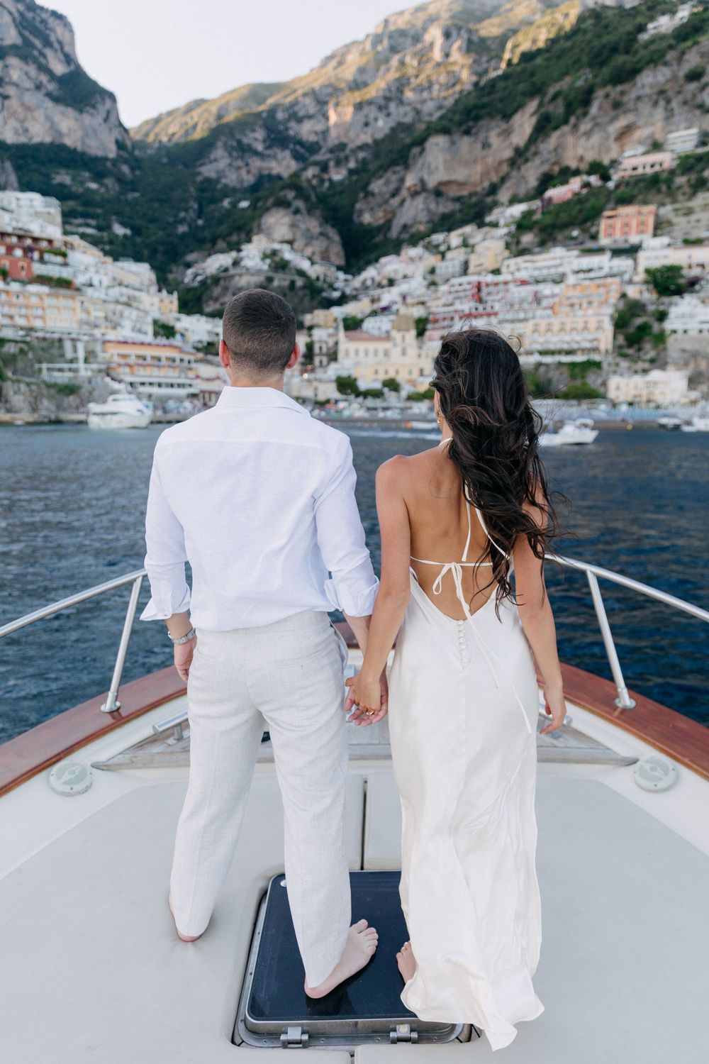 Couple in white attire holding hands on yacht bow with Positano cliffside village backdrop.