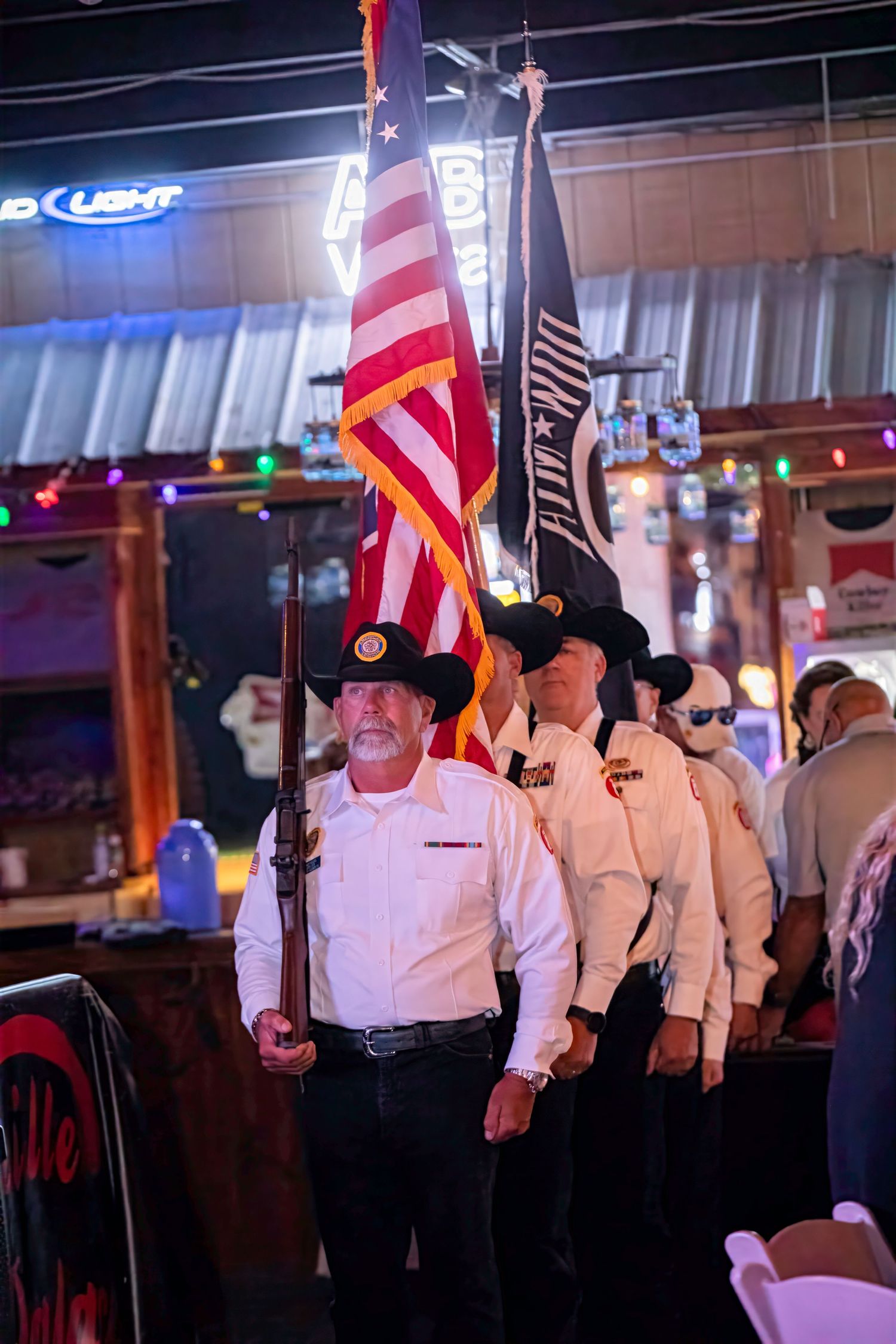 Color guard in black cowboy hats carrying American flags at an indoor venue with neon signs.