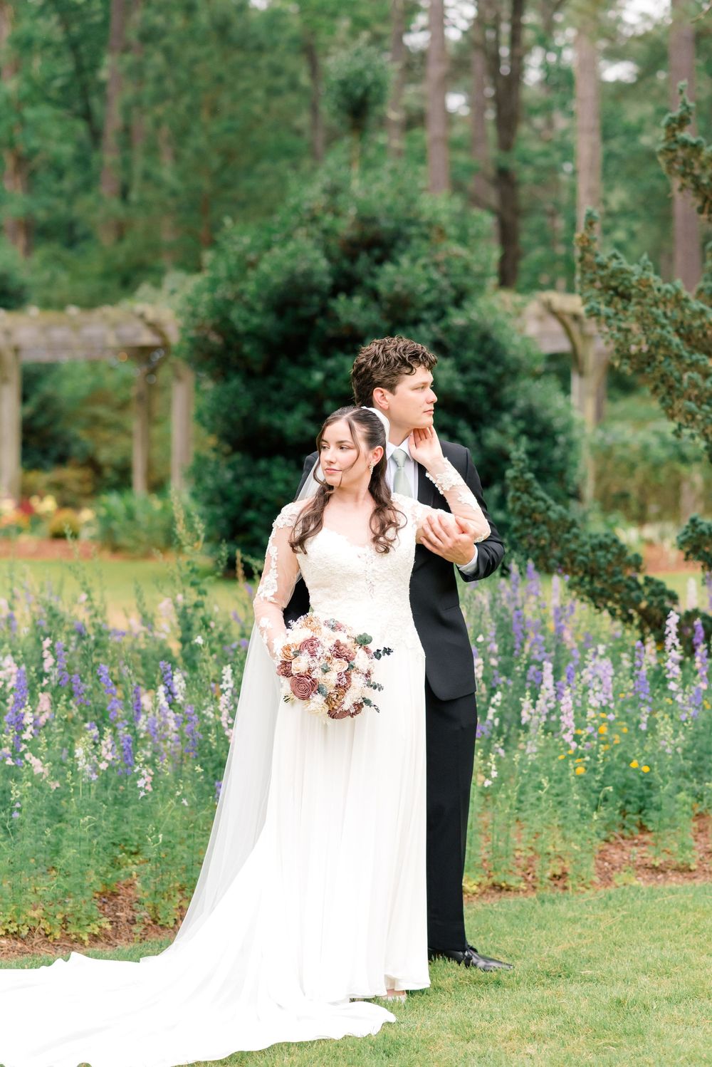 Bride and groom pose in a lush garden with purple flowers, she holds a bouquet in a white gown with veil.