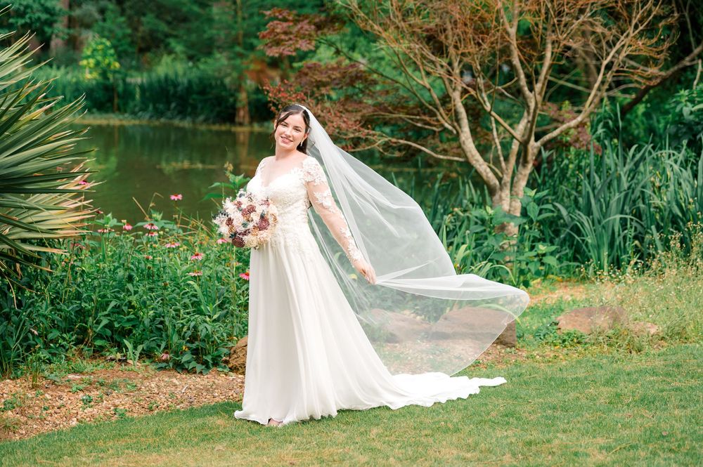 Bride in white gown and veil poses outdoors by a pond surrounded by lush greenery on her wedding day.