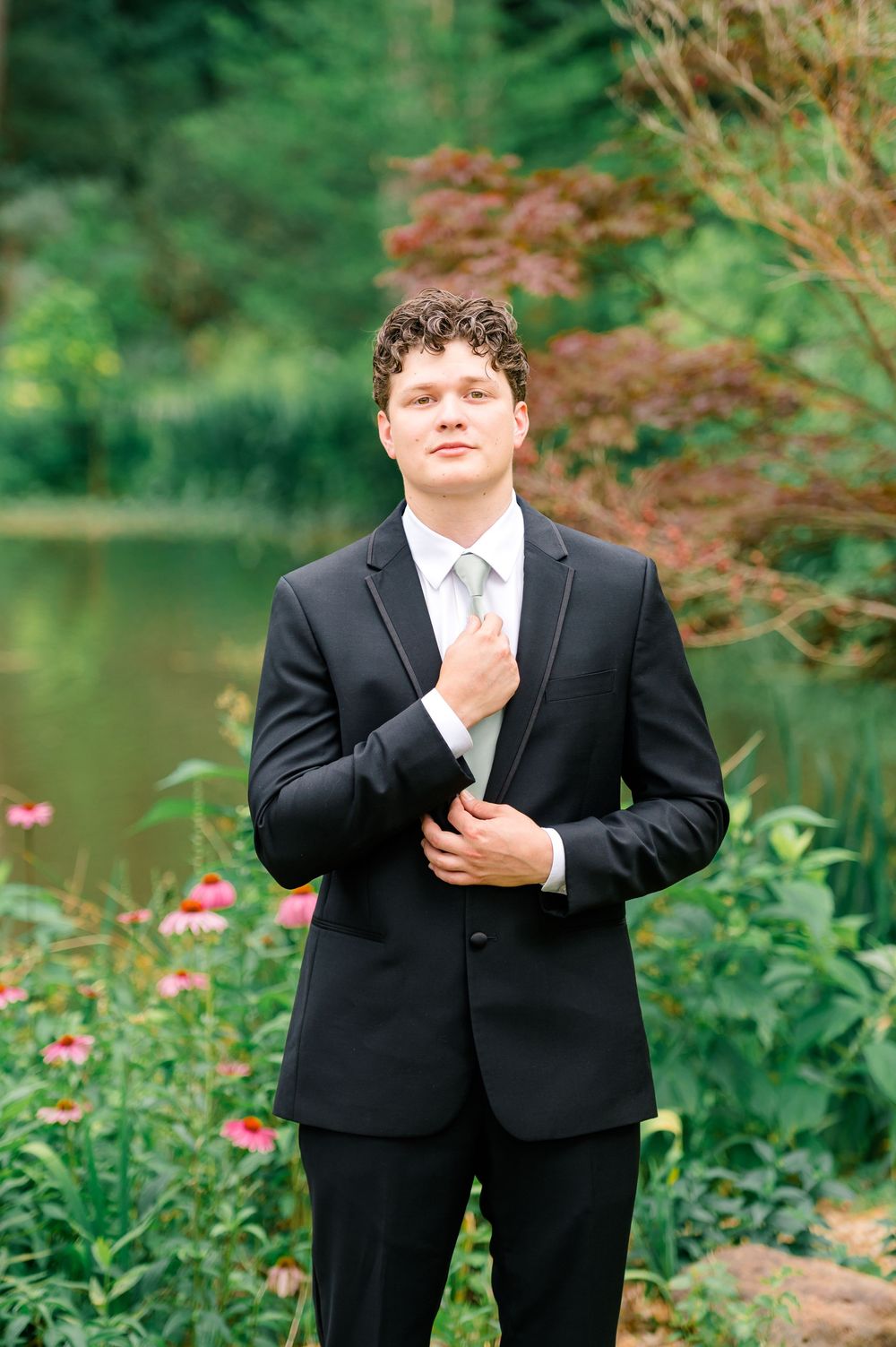 Groom in black suit adjusts his jacket near a serene pond surrounded by lush greenery and pink flowers.