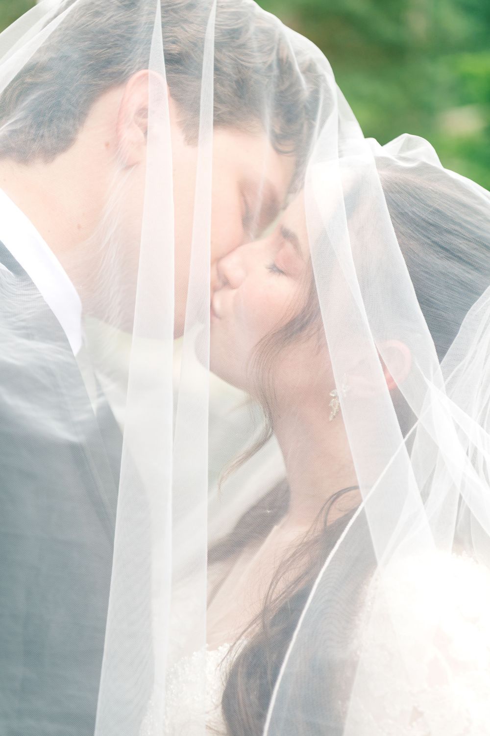 Bride and groom sharing a romantic kiss under a flowing white wedding veil outdoors.