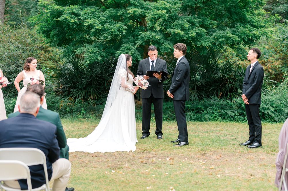 Outdoor wedding ceremony with bride in white gown and veil, groom in black suit, and officiant on green lawn.