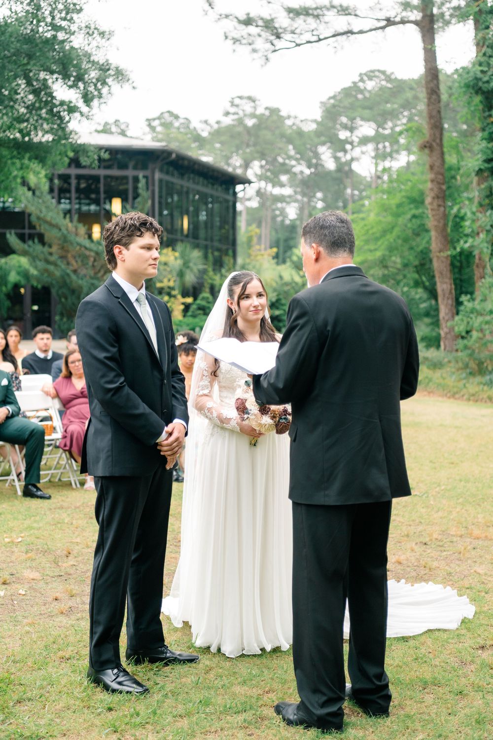 Outdoor wedding ceremony with bride in white gown and groom in black suit exchanging vows before officiant in wooded venue.
