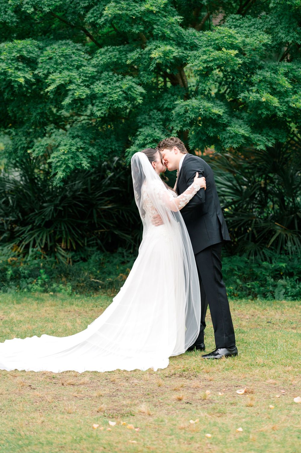 Bride and groom sharing a romantic forehead touch in lush garden, bride in flowing white gown with long veil.